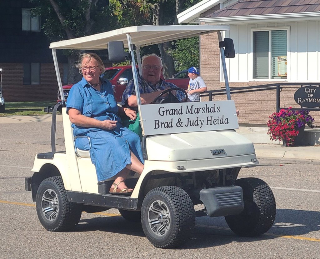 Grand Marshals Brad and Judy Heida riding in a golf cart during a parade in Raymond, featured on the Willmar Dental page