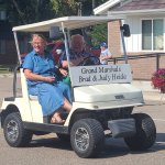 Grand Marshals Brad and Judy Heida riding in a golf cart during a parade in Raymond, featured on the Willmar Dental page